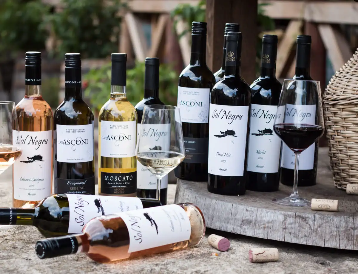 A variety of wine bottles and glasses from Asconi Winery displayed on a wooden table. The bottles feature labels with different wine names like Sol Negru, Pinot Noir, Merlot, Cabernet Sauvignon, Moscato, and Riesling. Some bottles are lying down, while others are standing upright. A wine glass is partially filled with red wine