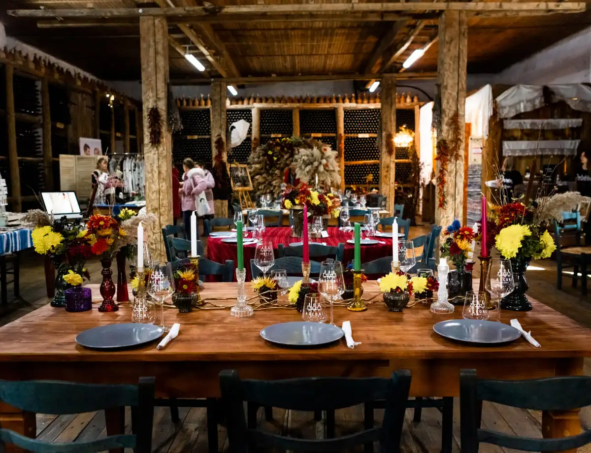 A rustic banquet hall in a winery with exposed wooden beams and brick walls. Tables are set with colorful floral arrangements, candles, and glassware. Guests can be seen in the background, and wine bottles are displayed on shelves