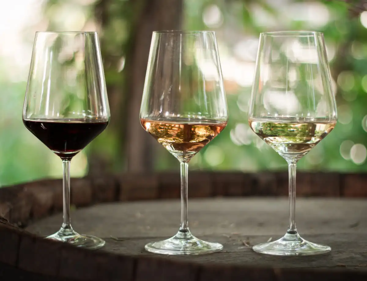 Three wine glasses, from left to right: a red wine glass, a rosé wine glass, and a white wine glass, sitting on a wooden barrel with a green and blurred background